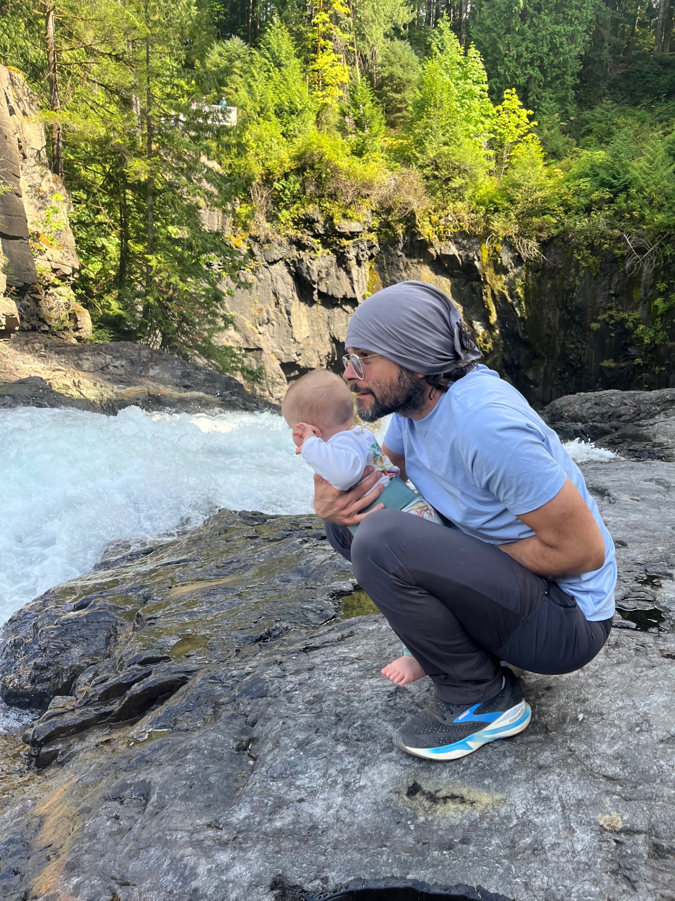 A father crouching with his baby on the rocks beside a river